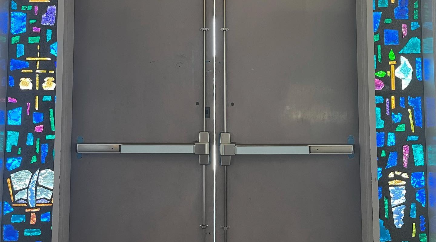 Panic bars installed on a set of doors in a church setting. Stained glass windows frame the doors on either side.